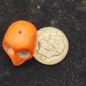 May include: A small orange skull bead next to a US dime coin with the year 1981.