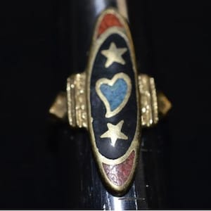 May include: A vintage ring with a gold-tone band and a central oval design. The design features a black background with a blue heart, two gold stars, and red and orange accents. The ring is a unique piece of jewelry.