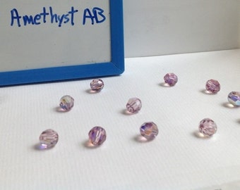 May include: A close-up of 11 light amethyst AB beads. The beads are round and have a faceted design. The beads are arranged in a scattered pattern on a white surface. The text "Light Amethyst AB" is written on a blue whiteboard in the background.