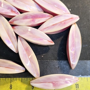 May include: A group of 11 pink and white iridescent shell beads in a leaf shape. The beads are approximately 1 inch long and are arranged in a pile on a black surface. A yellow ruler is visible in the bottom of the image.