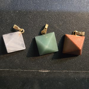 Puede incluir: Tres colgantes de cristal en forma de pirámide con un cierre de metal dorado. Los colgantes están hechos de cuarzo rosa, aventurina verde y jaspe rojo.