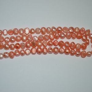 May include: A strand of peach-colored freshwater pearls. The pearls are round and have a slightly irregular shape. They are strung on a clear thread.