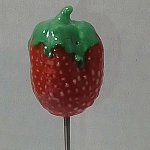 May include: A red strawberry-shaped decorative item with a green stem, mounted on a long, thin, silver-colored metal rod. The strawberry has a textured surface and is likely a garden ornament or decorative item.