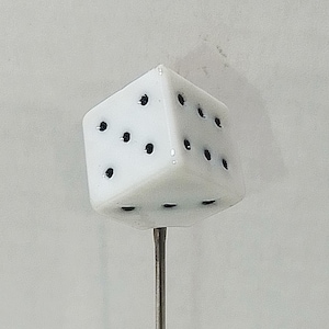 May include: A white dice with black dots, mounted on a thin, silver-colored metal rod. The dice is a cube shape, with each side displaying a different number of dots. The rod is centered beneath the dice.