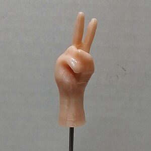 May include: A small, peach-colored hand figurine in a peace sign gesture, mounted on a thin, silver rod. The hand is detailed with visible knuckles and a smooth texture, set against a plain, light gray background.