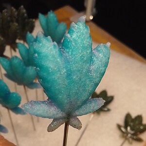 May include: Close-up of several teal-colored, sugar-coated flower-shaped decorations on thin metal wires. The flowers have a textured, crystalline appearance. Some are in focus, while others are blurred in the background. The background is a white surface.
