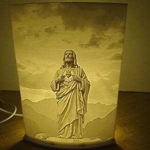May include: A beige, rectangular lamp featuring a detailed relief of Jesus Christ with a heart emblem. The lamp's design includes a landscape with mountains and clouds, illuminated from within, creating a warm glow.