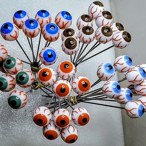 May include: A collection of glass eye pins with various iris colors, including blue, green, brown, and orange. Each eye is detailed with red veins and set on a silver pin. The pins are clustered together, creating a unique decorative display.