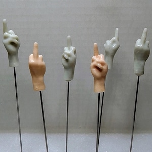 May include: A collection of five hand-shaped decorative items, each mounted on a thin metal rod. The hands are in various poses, including the middle finger gesture and the index finger pointing up. The hands are in shades of beige and off-white.