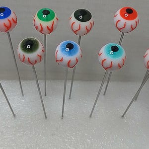 May include: A collection of ten eyeball-shaped pins with various iris colors, including blue, green, and orange. Each pin features a white sclera with red vein detailing and a silver pin stem. The pins are arranged in a white container.