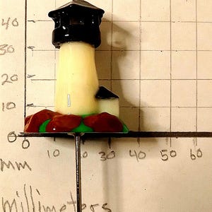 May include: A miniature lighthouse figurine with a black top, cream-colored tower, and a small adjacent building. The base features red and green accents. The lighthouse is mounted on a metal rod.