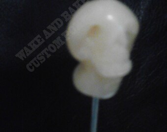 May include: A white sugar skull lollipop on a stick. The skull is detailed with a small yellow spot on the forehead.