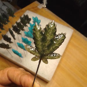 May include: A close-up of a green leaf-shaped decorative item on a wire stem. Other similar leaves in shades of green and blue are arranged on a white surface in the background. The leaf is detailed with a textured surface.