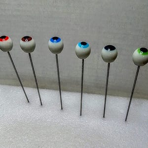 May include: Six novelty eye-shaped pins with different colored irises: red, brown, blue, light blue, black, and green. Each eye is mounted on a long, thin, silver-colored pin. The eyes are white with a black pupil.