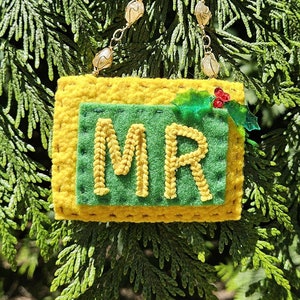 Könnte beinhalten: Handgefertigter gelb-grüner Schmuck mit den Buchstaben "MR". Der Schmuck ist mit einem kleinen Stechpalmenzweig verziert und hängt an einer Perlenkette. Der Hintergrund ist eine dichte Anordnung grüner Blätter.