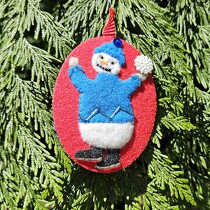 May include: A felt snowman Christmas decoration. The snowman is wearing a blue sweater, white trousers, and black boots. It is attached to a red oval with a red and white striped ribbon for hanging. The decoration is set against a backdrop of green pine needles.