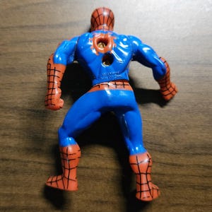 May include: A plastic Spider-Man action figure, viewed from the back, in a crouching pose. The figure is primarily blue with red accents on the arms, legs, and head. The figure has a hole in the back.