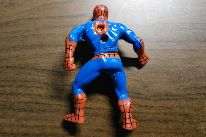 May include: A plastic Spider-Man action figure, viewed from the back, in a crouching pose. The figure is primarily blue with red accents on the arms, legs, and head. The figure has a hole in the back.