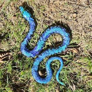 May include: A blue and teal snake ornament lies on grass and leaves. The snake has a detailed scale pattern, with colours blending from dark blue to teal. The snake is coiled and curved, with its head raised.
