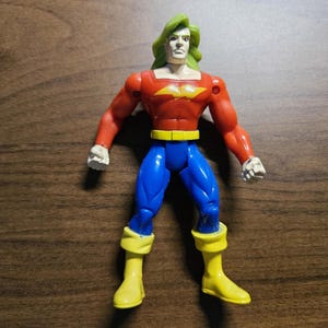 Könnte beinhalten: Eine Superhelden-Actionfigur mit rotem Oberkörper, blauer Hose und gelben Stiefeln. Die Figur hat grüne Haare und einen gelben Gürtel. Die Arme der Figur sind ausgestreckt, als ob sie bereit für eine Aktion wäre. Das Spielzeug ist aus Kunststoff.