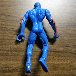 May include: A blue action figure with orange accents on the arms. The figure is posed with arms outstretched and legs slightly apart. The toy is made of plastic and has visible muscle definition.