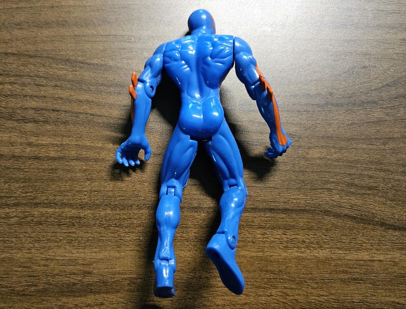 May include: A blue action figure with orange accents on the arms. The figure is posed with arms outstretched and legs slightly apart. The toy is made of plastic and has visible muscle definition.
