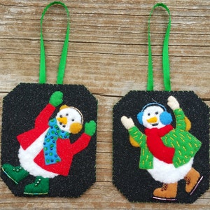 May include: Two handmade snowman decorations. Each decoration features a snowman with a white body, wearing a red coat, green mittens, and boots. One snowman has a blue scarf, the other a red scarf. Both have black backgrounds and green ribbon hangers.