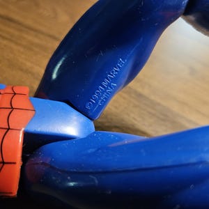 以下が含まれることがあります： 青と赤のアクションフィギュアのクローズアップ。フィギュアの腕は青色で、「&copy;1994 MARVEL CHINA」の文字がプリントされています。フィギュアの赤と黒のウェブデザインが見えます。