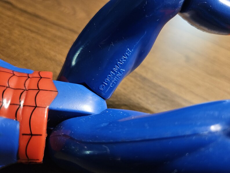以下が含まれることがあります： 青と赤のアクションフィギュアのクローズアップ。フィギュアの腕は青色で、「&copy;1994 MARVEL CHINA」の文字がプリントされています。フィギュアの赤と黒のウェブデザインが見えます。