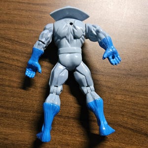 以下が含まれることがあります： 青い手袋とブーツがアクセントのグレーのアクションフィギュア。フィギュアは襟付きで、背面を視聴者に向けています。おもちゃはプラスチック製で、関節があります。