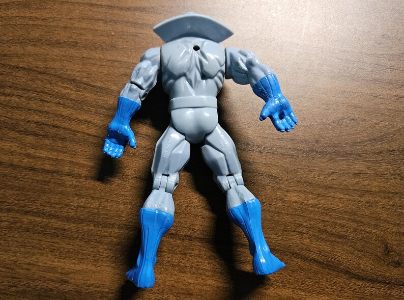 以下が含まれることがあります： 青い手袋とブーツがアクセントのグレーのアクションフィギュア。フィギュアは襟付きで、背面を視聴者に向けています。おもちゃはプラスチック製で、関節があります。