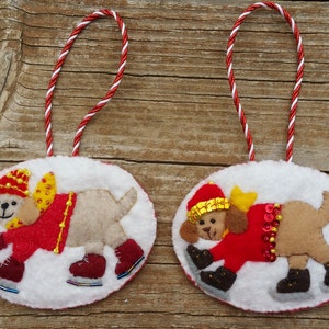 May include: Two oval Christmas tree decorations. Each features a felt dog wearing a red coat, hat, and ice skates. The dogs are set against a white background. The ornaments are suspended by red and white cord. The background is a wooden surface.