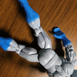 以下が含まれることがあります： 関節が可動するグレーとブルーのアクションフィギュア。フィギュアは青い手袋とブーツ、グレーの腕と胴体を持っています。フィギュアは木製の表面に横たわっています。