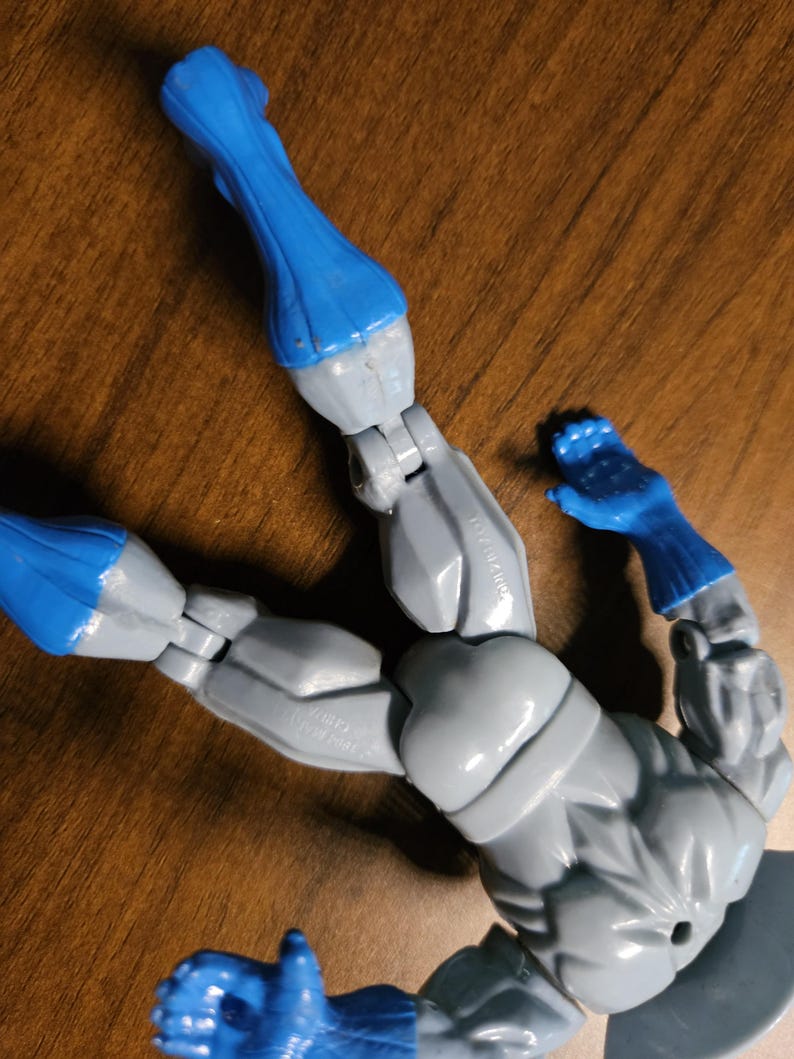 以下が含まれることがあります： 関節が可動するグレーとブルーのアクションフィギュア。フィギュアは青い手袋とブーツ、グレーの腕と胴体を持っています。フィギュアは木製の表面に横たわっています。