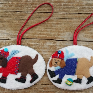 Könnte beinhalten: Zwei handgefertigte Hundeornamente aus Filz. Jedes Ornament zeigt einen braunen Hund in einem roten oder blauen Outfit mit passendem Hut. Die Ornamente sind weiß mit roten Aufhängeschnüren und mit blauen und goldenen Akzenten verziert.