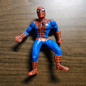May include: A Spider-Man action figure in a dynamic pose. The figure features a blue and red color scheme, with a detailed web pattern on the red sections. The figure is on a wooden surface.
