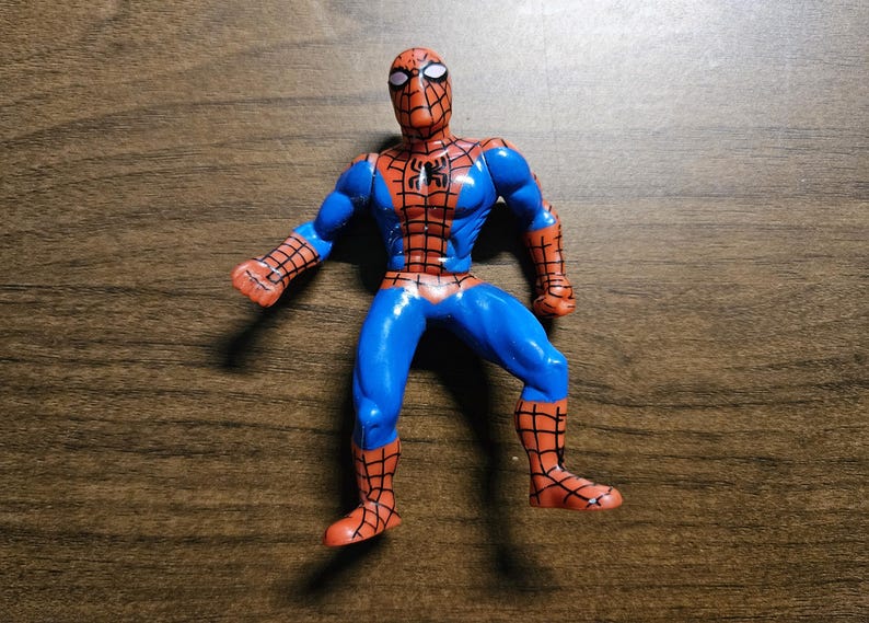 May include: A Spider-Man action figure in a dynamic pose. The figure features a blue and red color scheme, with a detailed web pattern on the red sections. The figure is on a wooden surface.