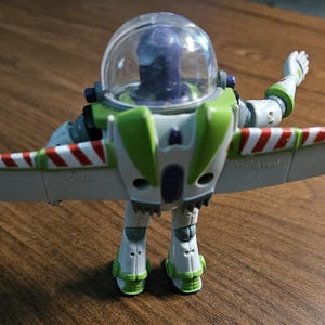 Puede incluir: Figura de acci&oacute;n de Buzz Lightyear, con casco transparente, traje espacial verde y blanco, y alas a rayas rojas y blancas. El juguete est&aacute; posando con un brazo levantado y las alas extendidas.