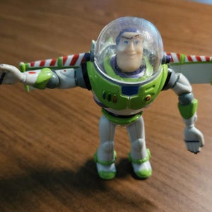 Puede incluir: Figura de acci&oacute;n de Buzz Lightyear en una pose din&aacute;mica. El juguete presenta un traje espacial verde y blanco con un casco transparente, alas y una cara sonriente. La figura est&aacute; de pie sobre una superficie de madera.