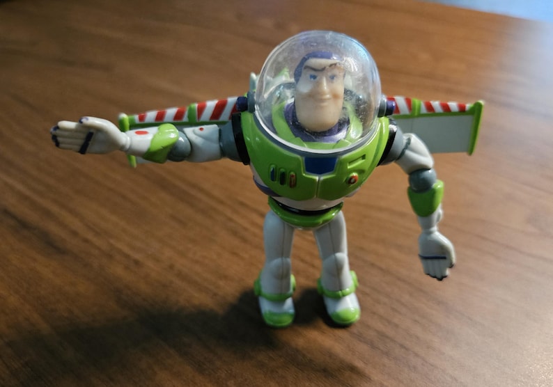 Puede incluir: Figura de acci&oacute;n de Buzz Lightyear en una pose din&aacute;mica. El juguete presenta un traje espacial verde y blanco con un casco transparente, alas y una cara sonriente. La figura est&aacute; de pie sobre una superficie de madera.