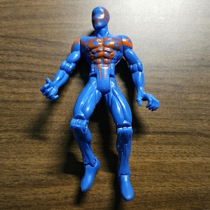 May include: A blue action figure with orange accents on the chest and shoulders. The figure is posed with arms outstretched and legs slightly bent. The toy is made of plastic and has a glossy finish.
