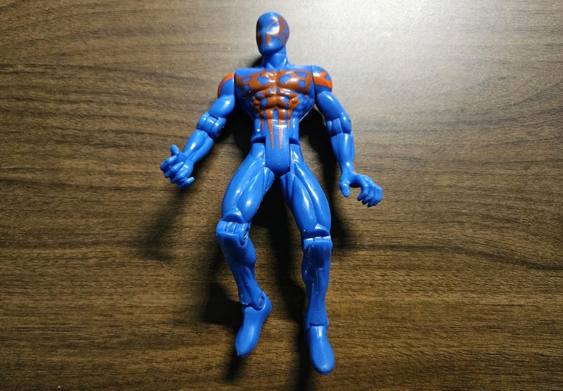 May include: A blue action figure with orange accents on the chest and shoulders. The figure is posed with arms outstretched and legs slightly bent. The toy is made of plastic and has a glossy finish.