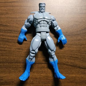 以下が含まれることがあります： 青い手袋とブーツを履いた灰色のフィギュア。このフィギュアは筋肉質な体格で、厳しい表情をしています。フィギュアは木製の表面に立っています。