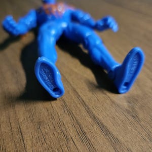 May include: A blue action figure of a superhero, lying on a wooden surface. The figure has red accents and is marked with text on the feet. The figure is made in China.