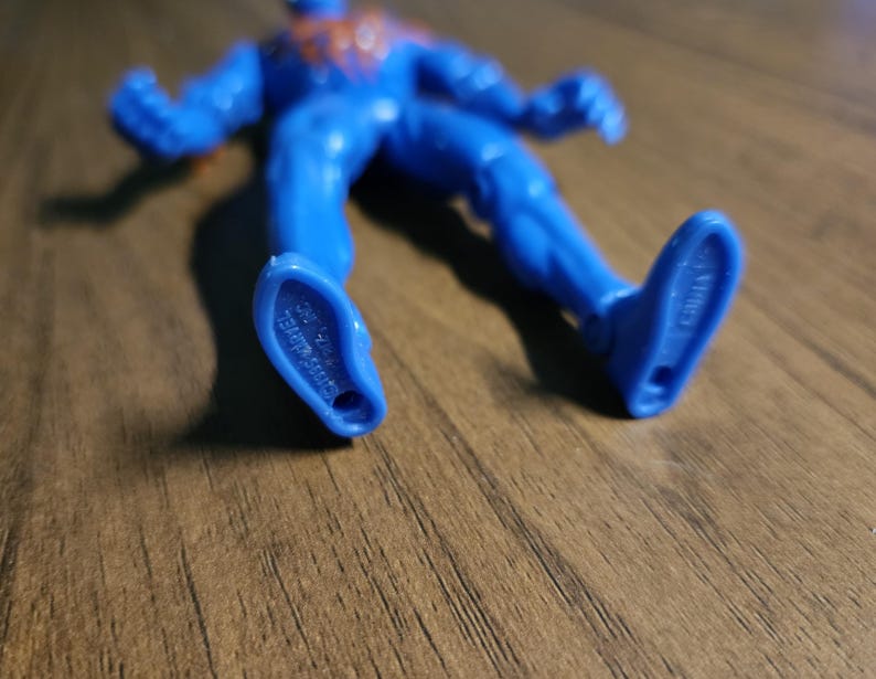 May include: A blue action figure of a superhero, lying on a wooden surface. The figure has red accents and is marked with text on the feet. The figure is made in China.