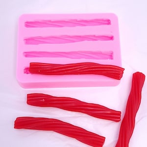 Puede incluir: Un molde de silicona rosa con cuatro cavidades con forma de regaliz retorcido. Se muestran cuatro caramelos de regaliz rojos junto al molde.
