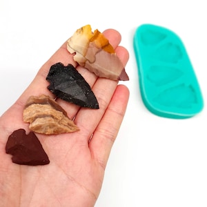May include: Four arrowhead-shaped pieces of stone, each with a different color and pattern, are displayed on a hand. The arrowheads are brown, black, tan, and red. A turquoise silicone mold with four arrowhead-shaped cavities is in the background.