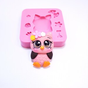 May include: A pink owl figurine with large, expressive eyes, a flower crown, and orange feet. A pink silicone mold with owl, flower, and bow shapes is in the background. The owl is likely a craft or baking decoration.