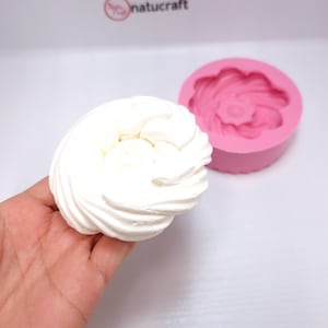 Op de afbeelding: Een witte, spiraalvormige lekkernij, vastgehouden in een hand, naast een roze siliconen mal in de vorm van een bloem. De afbeelding suggereert een bak- of knutselthema.