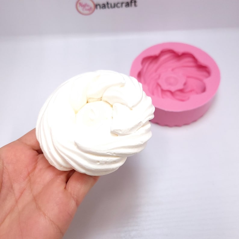 Op de afbeelding: Een witte, spiraalvormige lekkernij, vastgehouden in een hand, naast een roze siliconen mal in de vorm van een bloem. De afbeelding suggereert een bak- of knutselthema.